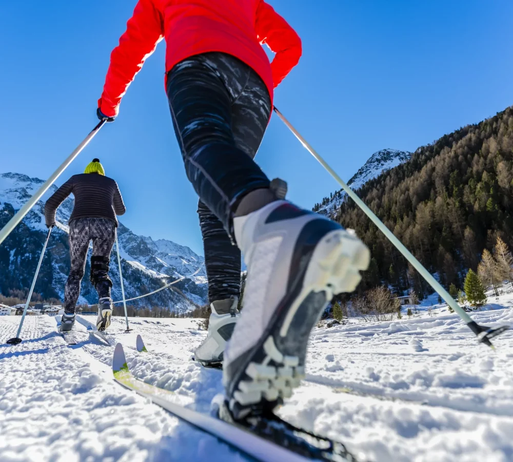 Due persone praticano sci di fondo su un sentiero innevato con montagne sullo sfondo.